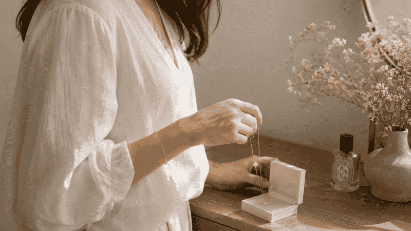 how to choose jewelry — woman selecting a delicate gold necklace from a jewelry box in morning light