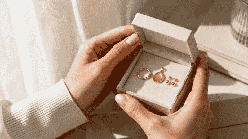 choosing jewelry metals — woman examining gold ring pendant and studs in open jewelry box by natural window light
