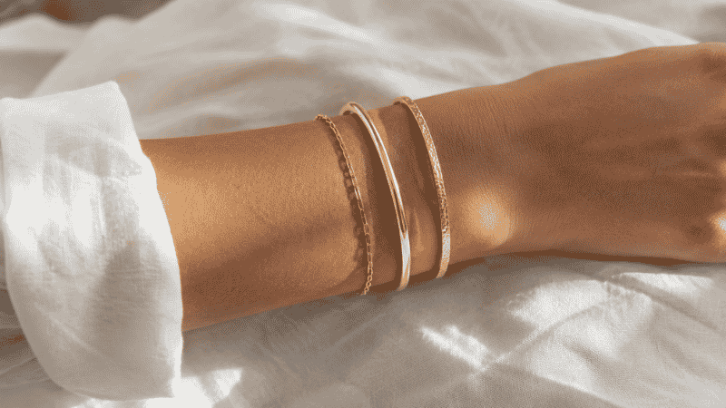 how to layer bracelets — three gold bracelets stacked on wrist showing thin chain bangle and textured cuff combination
