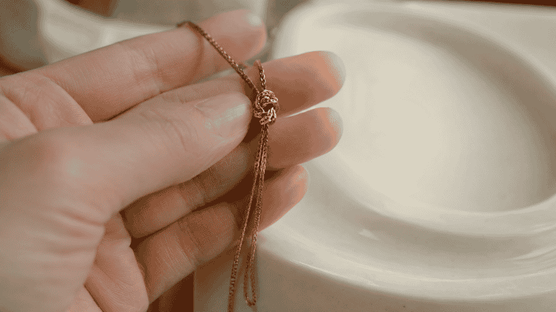 how to untangle a necklace — woman's hand holding a knotted delicate gold chain over white surface