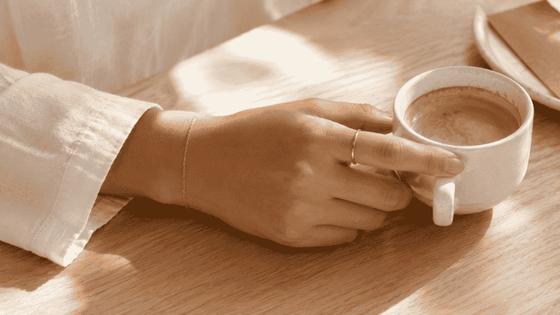 minimalist jewelry everyday wear — thin gold ring and delicate chain bracelet with morning coffee