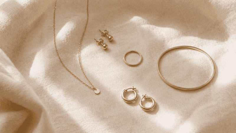 five minimalist jewelry essentials flat lay — gold studs, thin chain necklace, plain ring, slim bangle, and small hoops