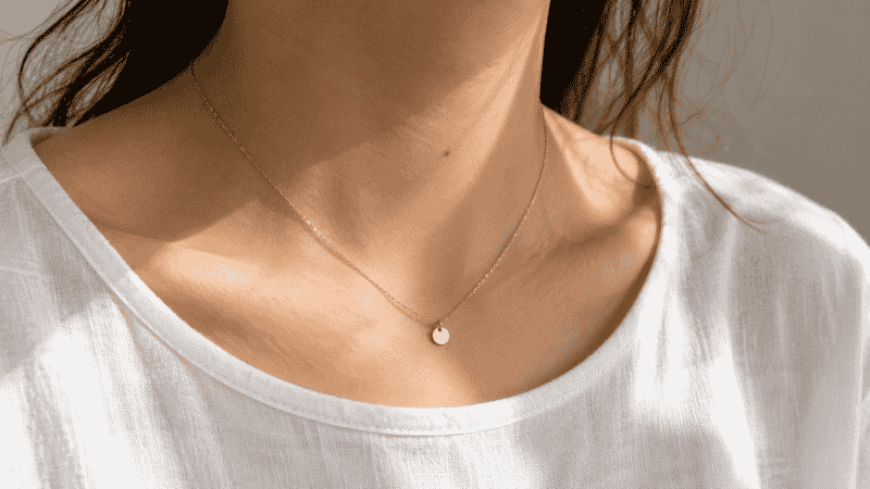 minimalist jewelry — delicate thin gold chain necklace with tiny disc pendant on collarbone with white linen top