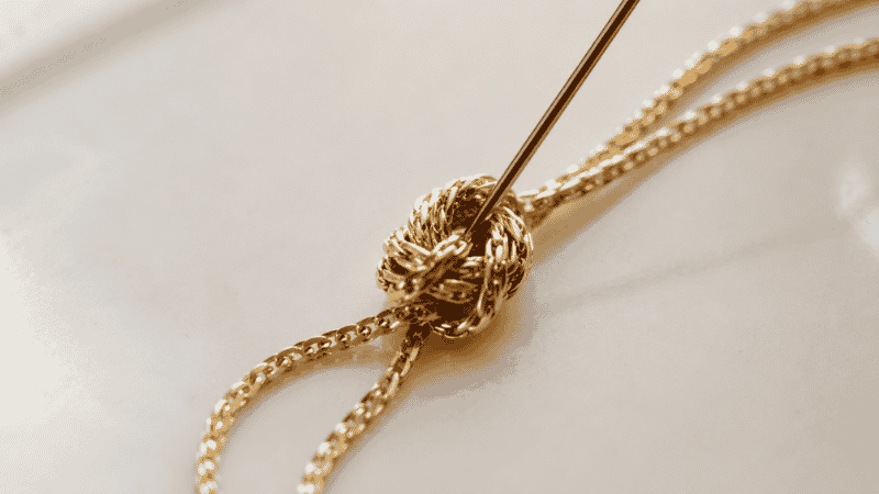 using a straight pin to untangle a necklace knot — pin inserted into center of gold chain knot on flat surface