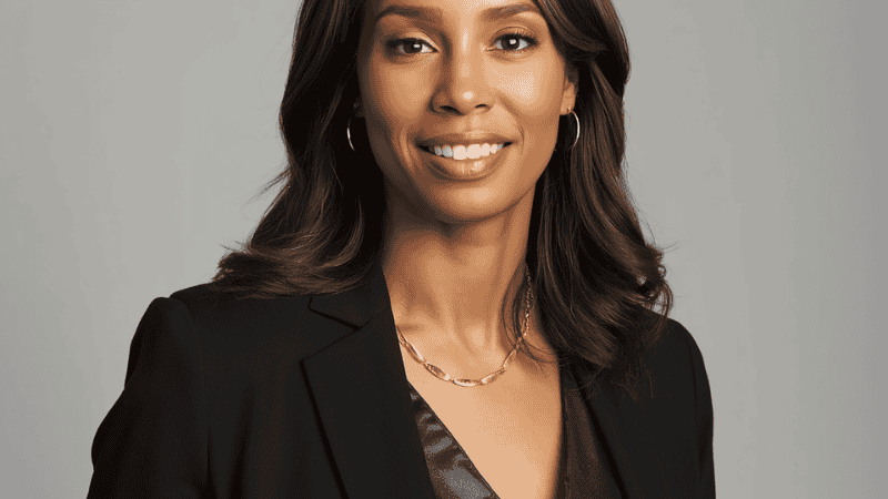 A professional woman wearing a structured navy blazer and white silk blouse, accessorized with a sleek 18k gold herringbone necklace that sits flat against the collarbone.