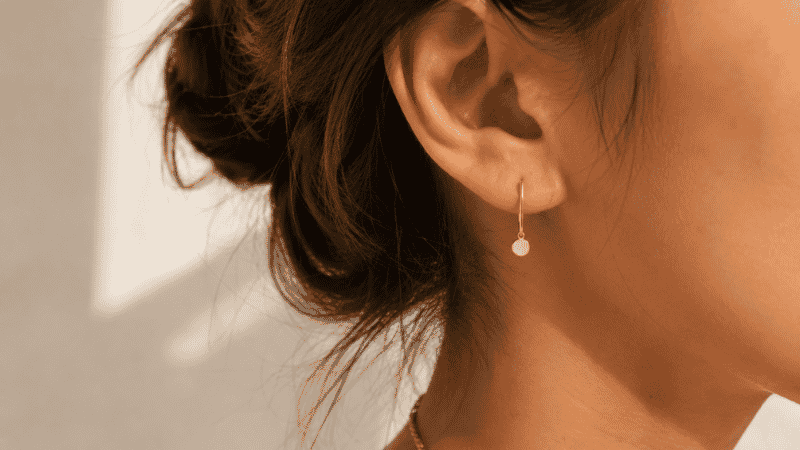 gold drop earrings — delicate bar with tiny disc pendant hanging just below lobe, hair up showing earring clearly