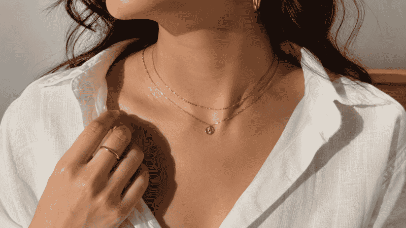 how to style jewelry — layered gold necklace stack with small hoops and thin ring, white linen shirt, editorial styling