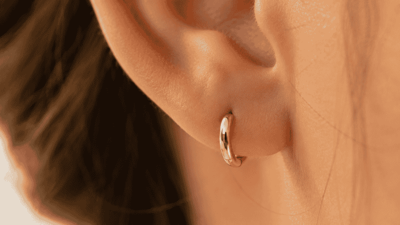 huggie earrings close-up — small gold huggie hoop sitting flush against earlobe showing the characteristic snug hugging fit