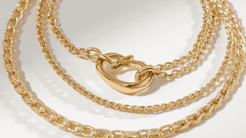 A detailed macro shot of a gold jewelry detangler clasp being used to organize and prevent tangling in a three-layer gold necklace stack at the back of the neck.