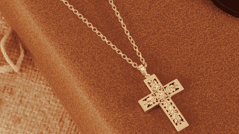 A flat-lay of a gold Kendra Scott cross necklace with pavé detailing, styled alongside a distressed leather journal and a pair of chic tortoiseshell sunglasses for a modern, grounded look.