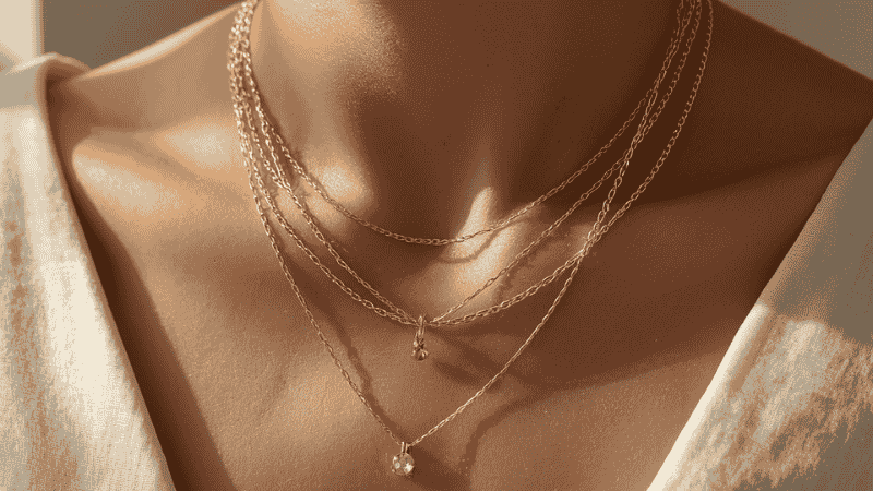 A sophisticated close-up of a woman's layered 18k gold necklace stack, featuring a dainty choker, a textured paperclip chain, and a medallion pendant against a neutral linen background.
