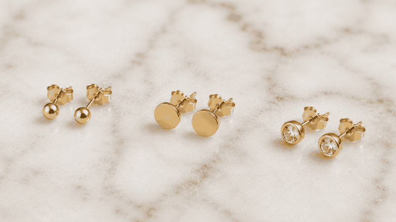 stud earrings types — three pairs of small gold studs including ball studs disc studs and bezel stone studs flat lay