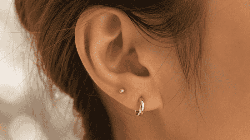 types of earrings — gold huggie earring on lobe with tiny gold stud on second piercing, minimal ear stack styling