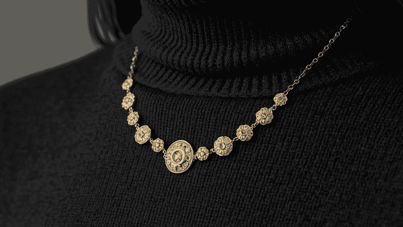 A professional close-up of a 10-motif Van Cleef Alhambra necklace in 18k Guilloché yellow gold layered over a dark charcoal cashmere turtleneck, showcasing the "V" focal point and textured gold shimmer