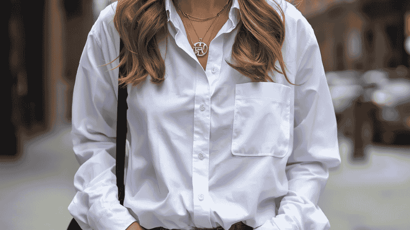 A lifestyle street-style shot of a woman wearing a gold Van Cleef Sweet Alhambra pendant layered with an unbranded gold chain, paired with an oversized white poplin shirt and distressed vintage denim